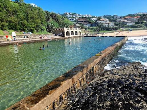 Swimmers in the Thompson's Bay tidal pool, with the historic beach building and the wall holding back the surf